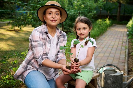 Pleasant young woman, mom and her daughter holding soil with growing cucumber seedlings in their hands, looking at camera. Family agribuisness, eco farming. Concept of World Environment Day. Ecology.