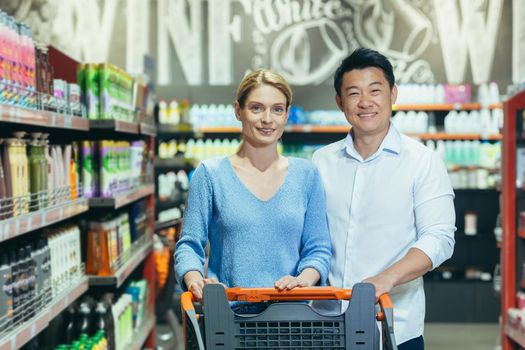Portrait of happy shoppers in supermarket, interracial couple Asian man and woman, smiling and looking at camera, among shelves with goods