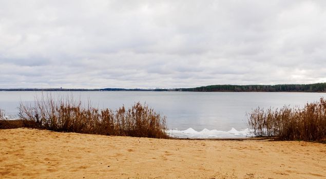 Calm lake, forest on the distant horizon, cloudy sky with clouds and sand on the shore.