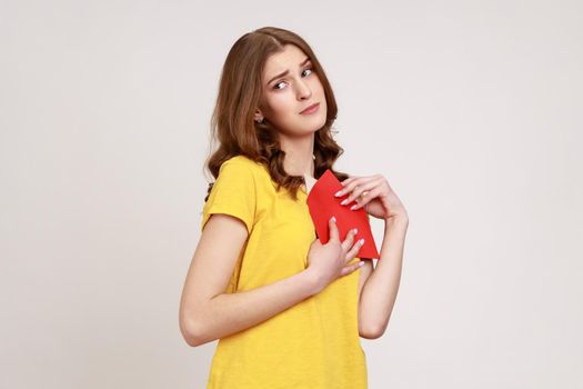 Love letter on Valentine's day. Portrait of teen girl in yellow casual T- shirt hugging letter in red envelope, holding greeting card with expression. Indoor studio shot isolated on gray background.