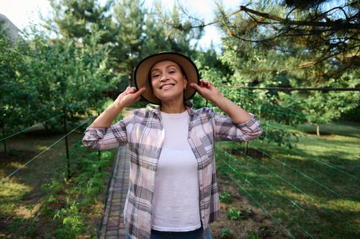 Portrait of young cheerful woman, farmer, wearing a straw hat and plaid shirt, smiling toothy smile, enjoying gardening on a a beautiful sunny summer day. Organic farm, growth of eco-friendly products
