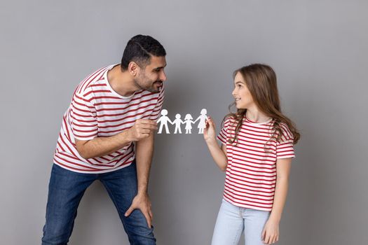 Portrait of happy delighted father and daughter in striped T-shirts, holding paper people chain, family relationship, childhood and parenthood. Indoor studio shot isolated on gray background.
