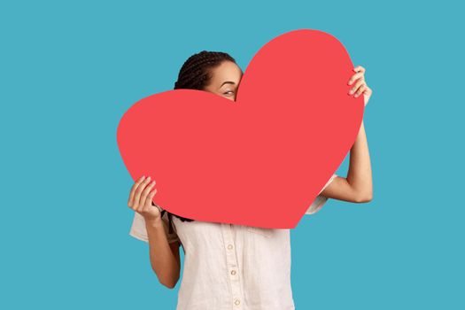 Portrait of unknown woman holding big red heart in hands, peeping from love symbol, being shy to tell about her feelings, wearing white shirt. Indoor studio shot isolated on blue background.