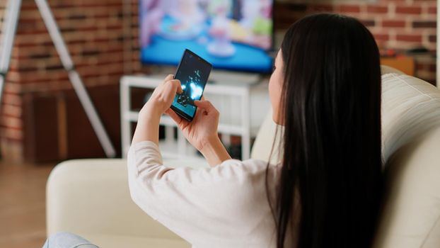 Playful woman working remotely while playing space shooter game on smartphone at home. Childish asian person enjoying mobile gaming while sitting on sofa in modern apartment.