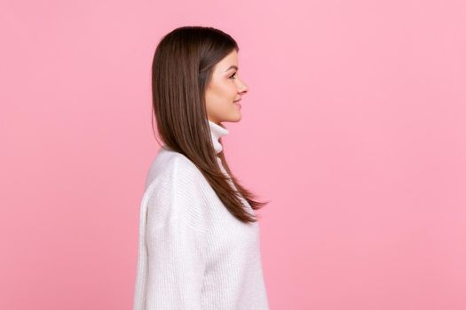 Side view portrait of smiling brunette female looks ahead, expressing happiness, being in good mood, wearing white casual style sweater. Indoor studio shot isolated on pink background.