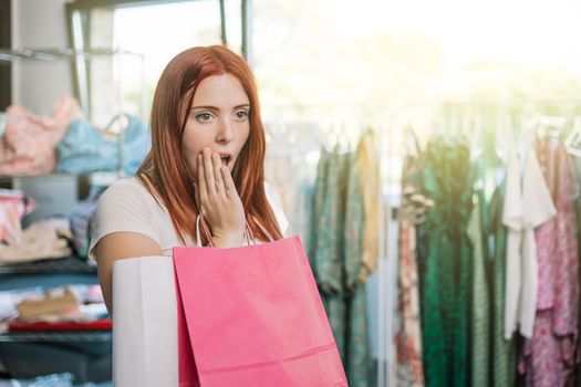 young red-haired girl surprised and excited, by the clothes she is looking at in the window, shopping in a small clothing shop. girl spending time shopping. concept of shopping. natural light from the window of the window.