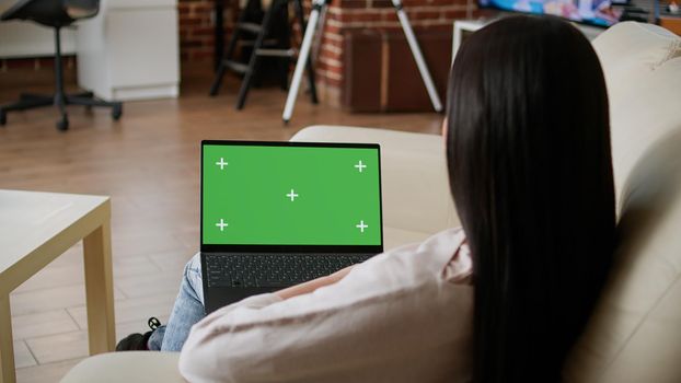 Woman sitting on couch inside apartment with portable computer having green screen display. Young adult person sitting on sofa at home while looking at laptop having chroma key isolated background.