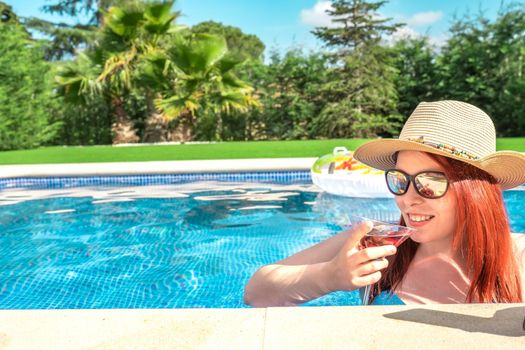 young woman with hat and sunglasses, having a refreshing drink inside the swimming pool. young girl on summer holiday sunbathing by the pool. concept of summer and free time. outdoor garden, natural sunlight.