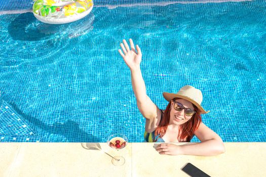 young woman with red hair smiling, waving from inside the pool, cooling off on a sunny day. young girl on summer holiday sunbathing by the pool. concept of summer and leisure time. outdoor garden, natural sunlight.