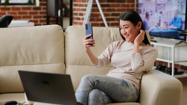 Smiling heartily woman taking selfie photos with smartphone while working remotely from home. Joyful person photographing herself with modern touchscreen phone device while doing remote work