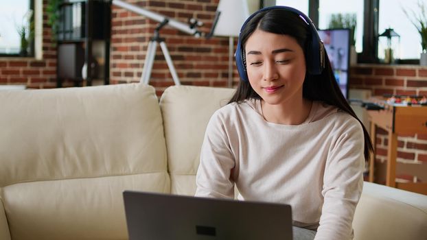 Cheerful woman wearing wireless headphones while working remotely at home on laptop. Happy smiling heartily young adult person doing remote work on modern computer while listening to music.