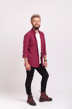 Full length body of happy bearded man in stylish plaid shirt and casual black pants smiling looking at camera, hipster male rejoicing. Indoor studio shot isolated on gray background