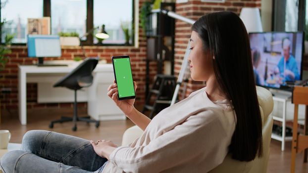 Happy smiling heartily woman having smartphone with green screen display while sitting at home on sofa. Joyful person sitting on couch with mobile cellphone having mockup chroma key background.
