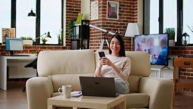 Attractive woman chatting on smartphone while sitting on sofa at home and smiling heartily at camera. Happy young adult person sending messages on modern mobile phone while working remotely.