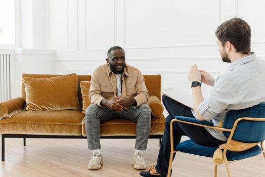 The therapist takes notes while talking to a depressed male patient and talks to an African American patient. Desperate black man suffering from nervous breakdown or depression having a session with a psychologist.