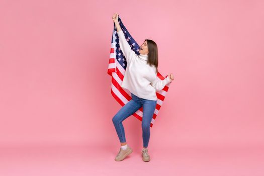 Full length photo of woman raised arms, holding american flag, celebrating national holiday, dancing, wearing white casual style sweater. Indoor studio shot isolated on pink background.