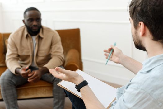 The therapist takes notes while talking to a depressed male patient and talks to an African American patient. Desperate black man suffering from nervous breakdown or depression having a session with a psychologist.