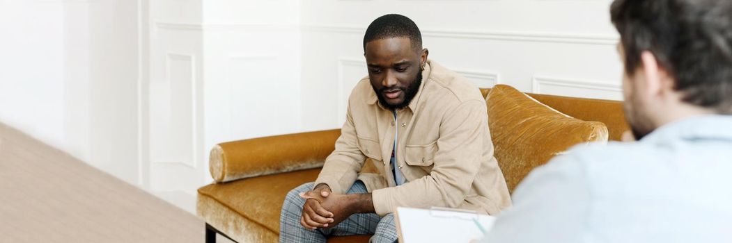 Unhappy young black man gay having session with professional psychologist at mental health clinic. Professional psychological help concept. Web banner.