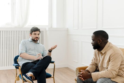 A male psychologist conducts psychological therapy with an African American man, makes notes in a clipboard