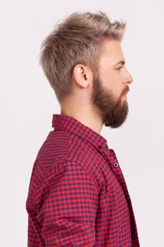 Profile portrait of confident bearded male with blond hair wearing red plaid shirt looking to side space with serious attentive face. Indoor studio shot isolated on gray background