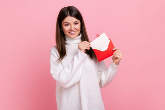Portrait of woman standing, holding envelope and looking at camera with toothy smile and enjoying, wearing white casual style sweater. Indoor studio shot isolated on pink background.