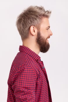 Profile portrait of happy bearded millennial man in red t-shirt sincerely smiling and looking to side, positive mood and optimistic lifestyle. Indoor studio shot isolated on gray background