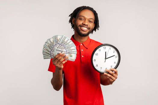 Happy young adult attractive man with dreadlocks wearing red casual style T-shirt, showing dollar cash and wall clock on camera, hourly payment. Indoor studio shot isolated on gray background.