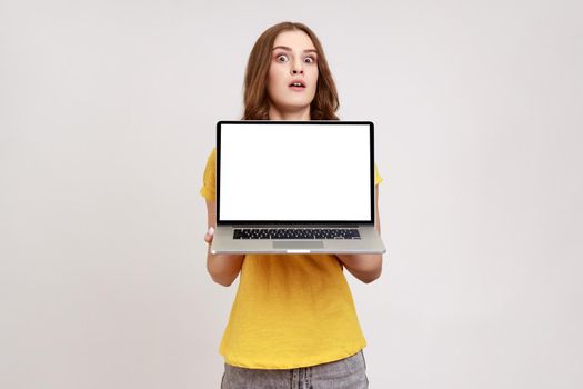 Portrait of surprised brown haired woman of young age in yellow T-shirt showing empty laptop screen, blank display for web advertising. Indoor studio shot isolated on gray background.