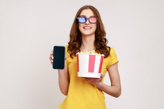 Image of beautiful young woman holding popcorn in bucket, showing smartphone with blank empty screen, smiling to camera, wears yellow T-shirt. Indoor studio shot isolated on gray background.