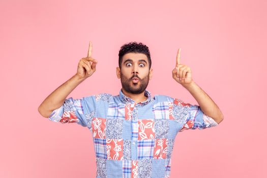 Attractive bearded man with astonished shocked facial expression wearing blue casual style shirt pointing up with both fingers, presenting copy space. Indoor studio shot isolated on pink background.