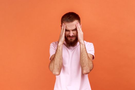Portrait of unhealthy sick young adult bearded man massaging temples, suffering from headache, migraine, wearing pink T-shirt. Indoor studio shot isolated on orange background.
