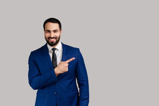 Positive bearded man pointing finger away with toothy smile on face, empty space for your advertisement, wearing official style suit. Indoor studio shot isolated on gray background.
