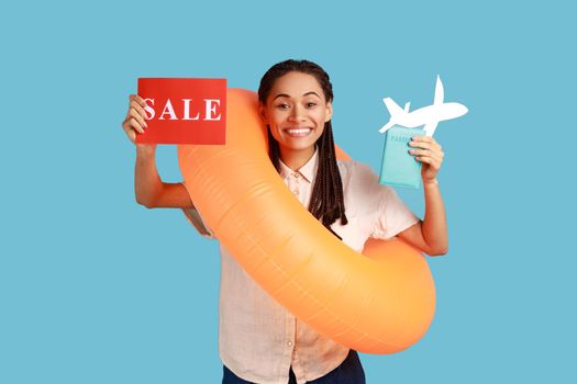 Portrait of joyful woman with rubber ring, holding card with sale inscription, and looking with toothy smile, enjoying summer sales for tours. Indoor studio shot isolated on blue background.