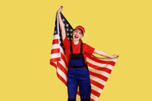 Excited worker woman standing with american flag in hands and yelling happily, celebrating holiday, wearing work uniform and red cap. Indoor studio shot isolated on yellow background.
