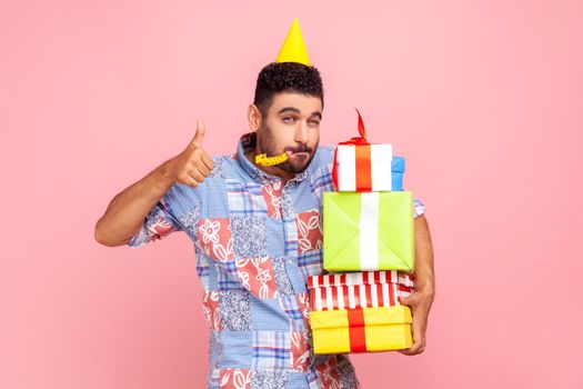 Portrait of bearded man with festive mood wearing party cone and casual shirt, showing thumb up to camera, holding stack of presents, and blowing horn. Indoor studio shot isolated on pink background.
