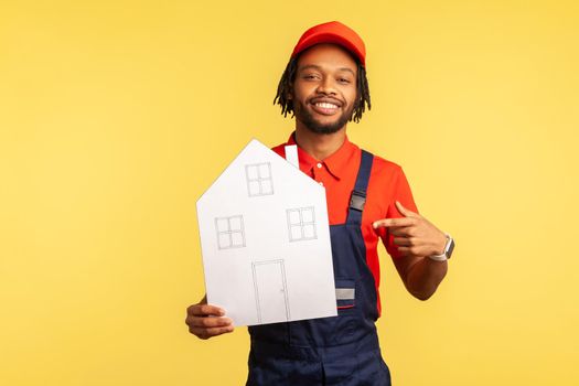 Smiling bearded handyman wearing red T-shirt and blue overalls, pointing finger at paper house in his hand, looking at camera with toothy smile. Indoor studio shot isolated on yellow background.