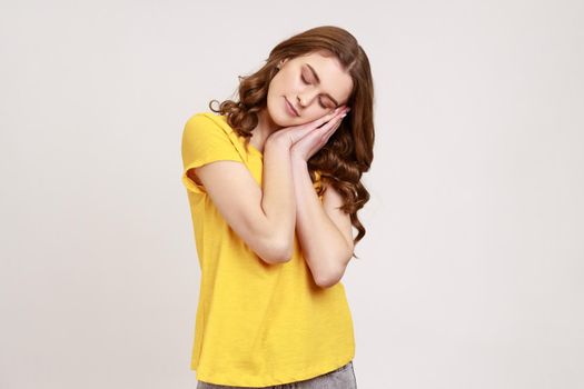 I want to sleep. Portrait of teenager girl with brown curly hair with closed eyes standing with arms on her cheeks like pillow and sleep. Indoor studio shot isolated on gray background.