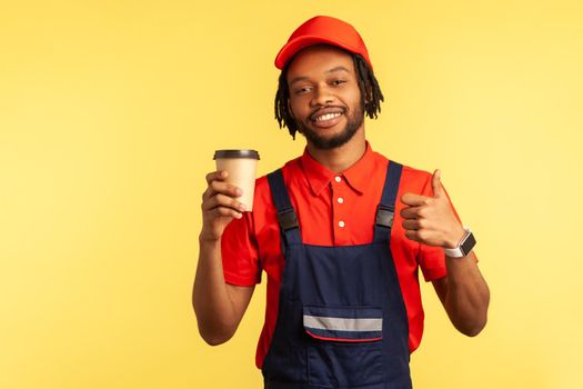 Portrait of smiling handyman wearing red T-shirt and blue overalls, holding coffee to go in hands and showing thumbs up, looking at camera. Indoor studio shot isolated on yellow background.