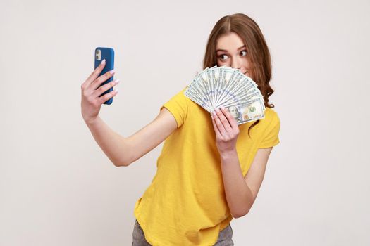 Portrait of rich teen girl with brown hair in T-shirt holding money and taking selfie by mobile phone, boasting of big profit, successful online betting. Indoor studio shot isolated on gray background