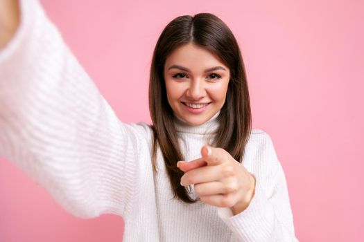 Happy brunette young adult smiling female making point of view photo, pointing finger to camera, wearing white casual style sweater. Indoor studio shot isolated on pink background.