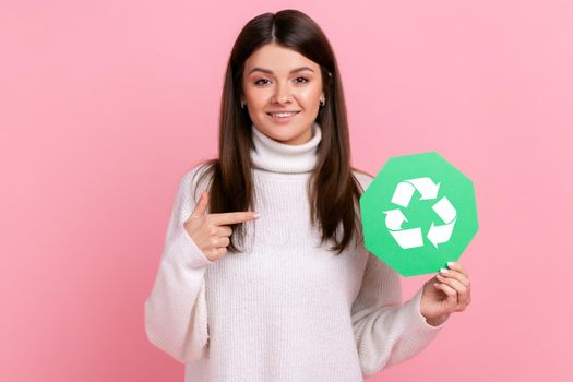 Satisfied female holding pointing at green recycling sign, looking at camera with charming smile, wearing white casual style sweater. Indoor studio shot isolated on pink background.