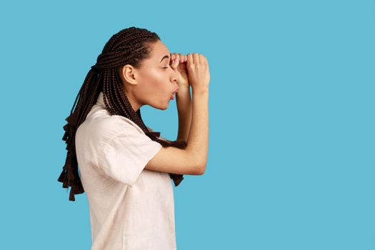 Side view of astonished woman with dreadlocks making glasses shape, looking through binoculars gesture with surprised expression, wearing white shirt. Indoor studio shot isolated on blue background.