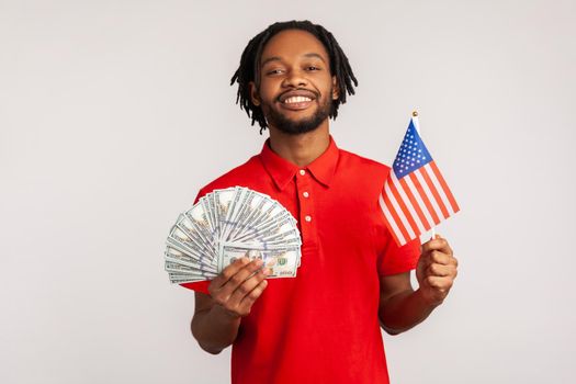 Portrait of smiling satisfied man with dreadlocks wearing red casual style T-shirt, holding american flag and dollars banknotes, celebrating success. Indoor studio shot isolated on gray background.