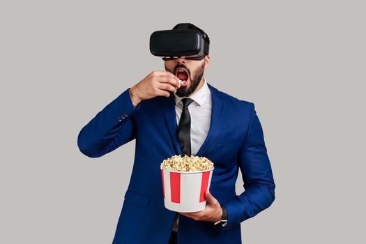 Portrait of excited amazed bearded businessman in VR headset watching movie with popcorn, eating tasty snack, wearing official style suit. Indoor studio shot isolated on gray background.