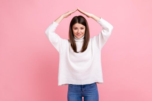 Woman making roof of hands above head looking at camera with toothy smile, feeling herself in safety, wearing white casual style sweater. Indoor studio shot isolated on pink background.