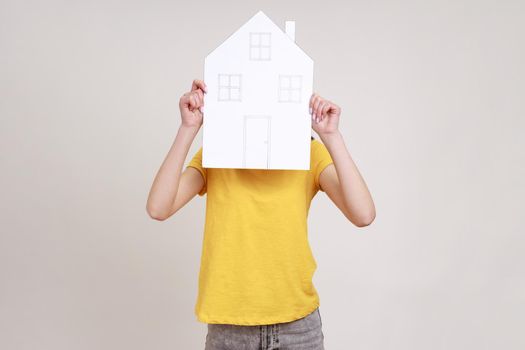 Unknown female in yellow T-shirt hiding her face behind paper house with drawn windows and door, government housing program, dreaming of new house. Indoor studio shot isolated on gray background.