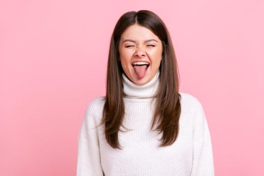Childish woman with dark hair showing tongue out, expressing positive emotions, having playful mood, wearing white casual style sweater. Indoor studio shot isolated on pink background.