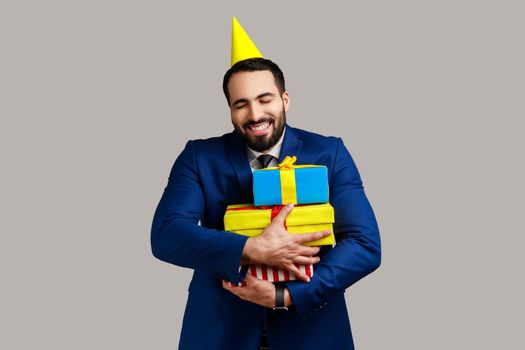 Extremely happy bearded man posing in party cone, holding embracing stack of presents, happy birthday, wearing official style suit. Indoor studio shot isolated on gray background.