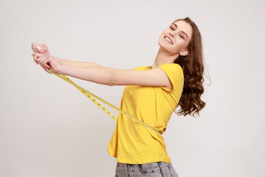 Profile portrait happy woman of toothy smile with toothy smile in yellow T-shirt measuring her waist with tape measure, satisfied with result. Indoor studio shot isolated on gray background.