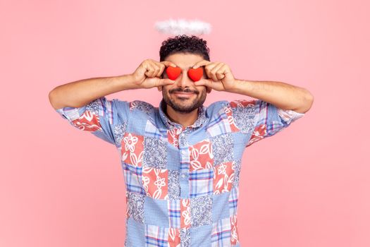 Portrait of young adult smiling man covering his eyes with red hears, wearing blue casual style shirt, having angelic nimb over head. Indoor studio shot isolated on pink background.